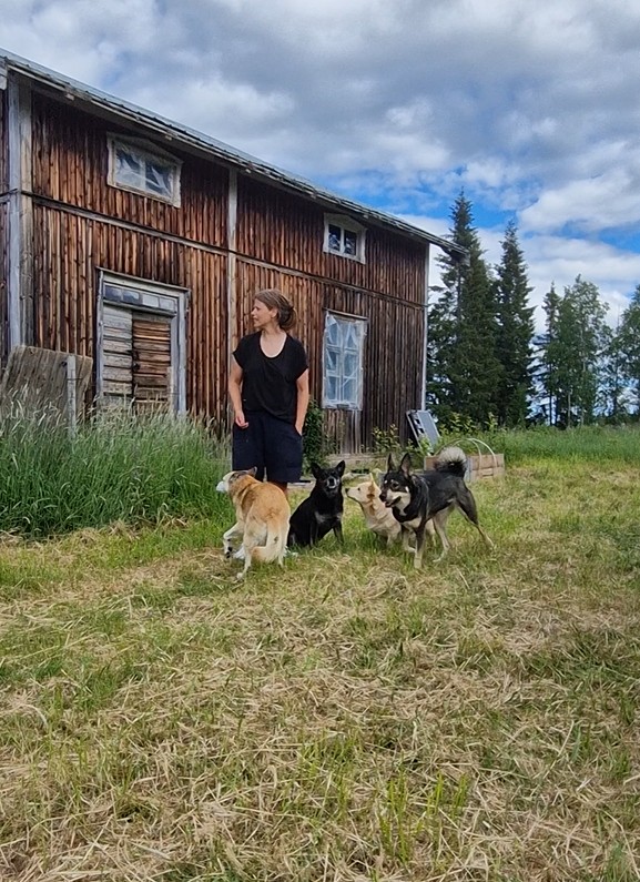 Jannice står utanför ett gammalt trähus med flera hundar runt sig.